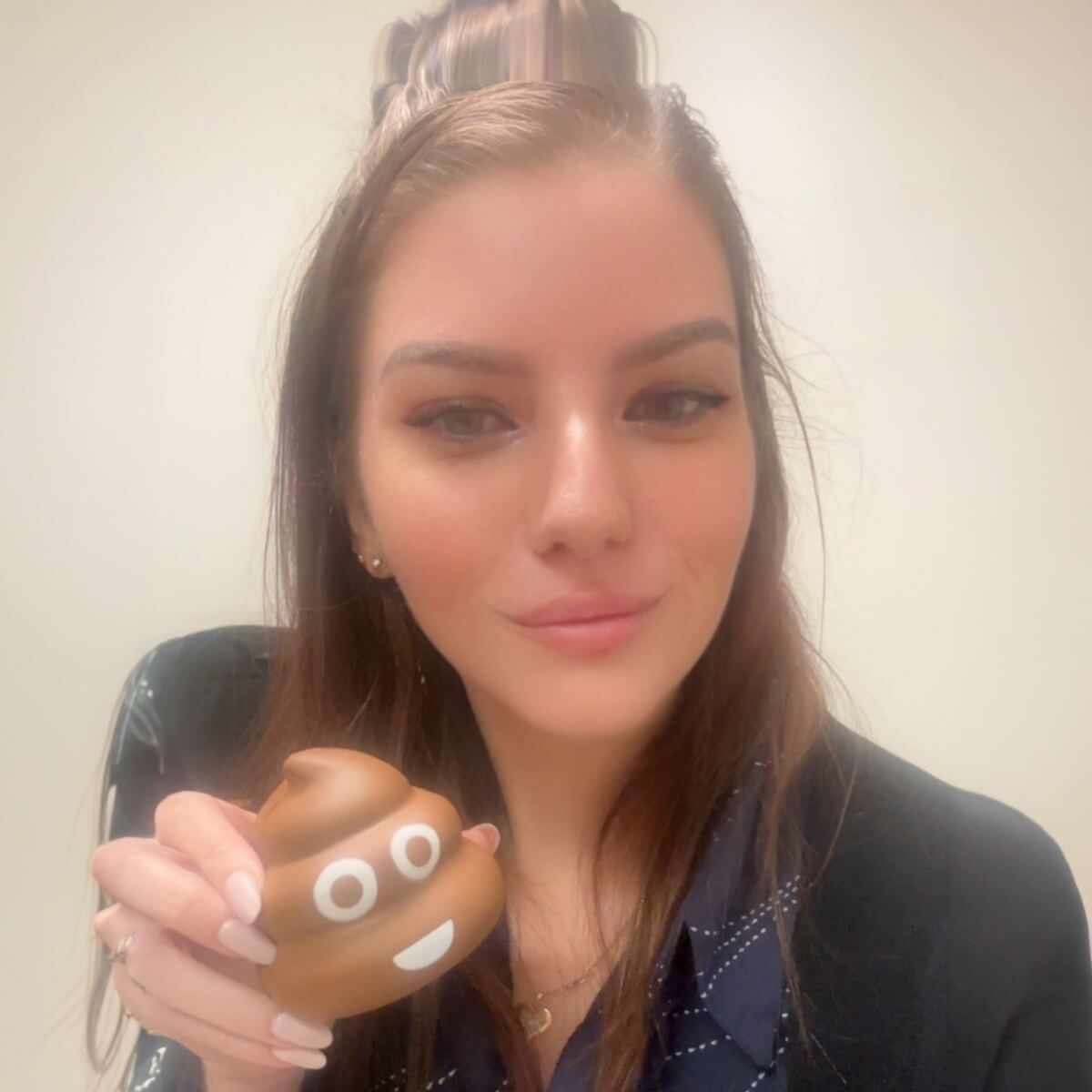 A woman holds a smiling poop emoji toy, smiling at the camera against a neutral background.