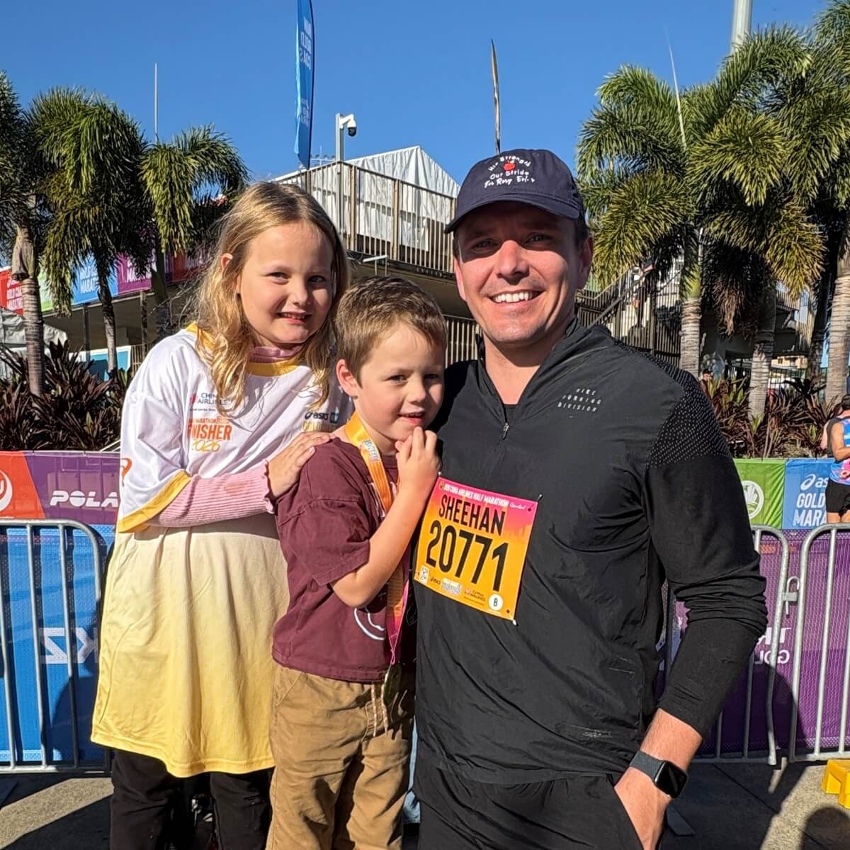 A man poses with two children at a race event, one wearing a finisher shirt and the other holding a medal.