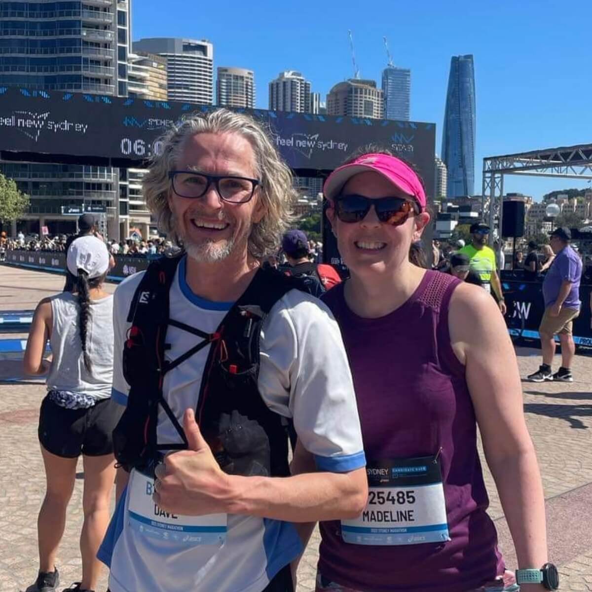 Two runners, a man with gray hair and a woman in a purple tank top, smile at a race event with city buildings in the background.