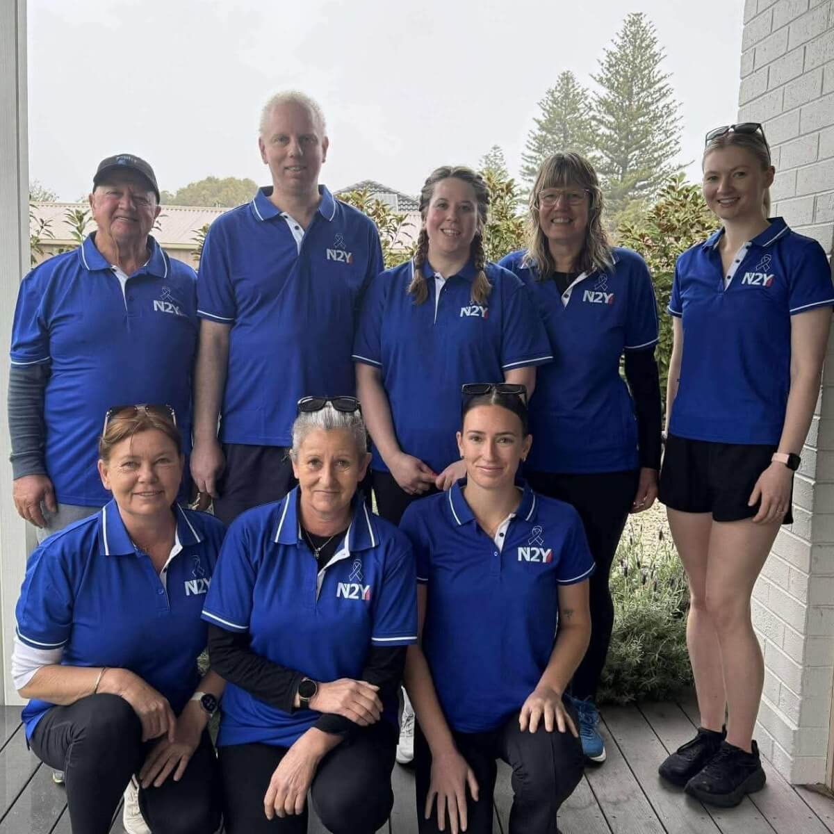 A group of eight people in blue shirts with "N2Y" logo, standing and sitting on a porch with greenery in the background.