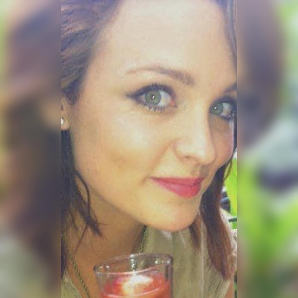 A close-up of a young woman with green eyes, smiling and holding a glass with a drink.