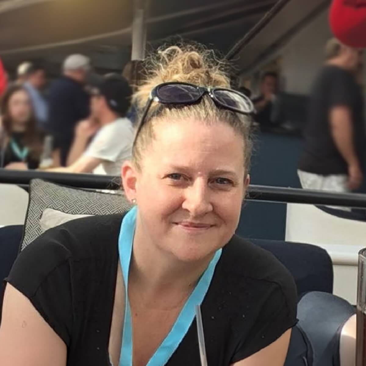 A woman with light brown hair styled in a bun, wearing a black top and a blue lanyard, smiles while seated outdoors.