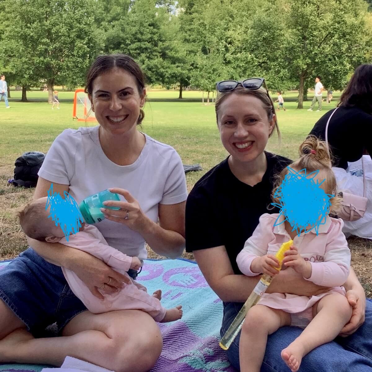 Two women sit on a blanket in a park, one feeding a baby and the other holding a toddler with a toy.