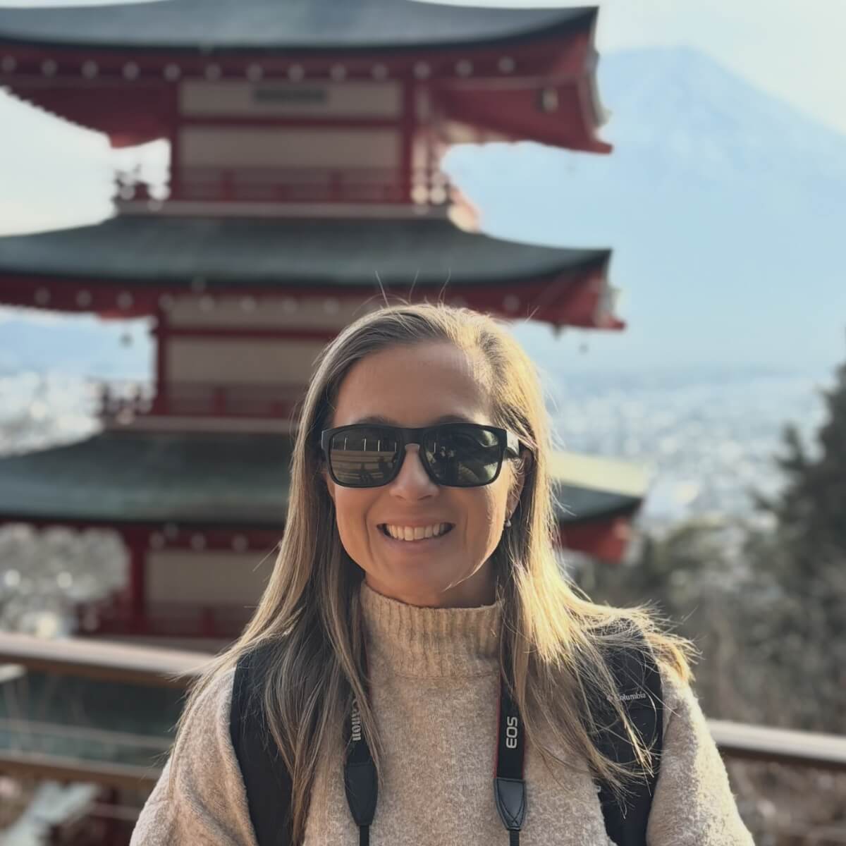 A woman in sunglasses smiles in front of a red pagoda with mountains in the background.