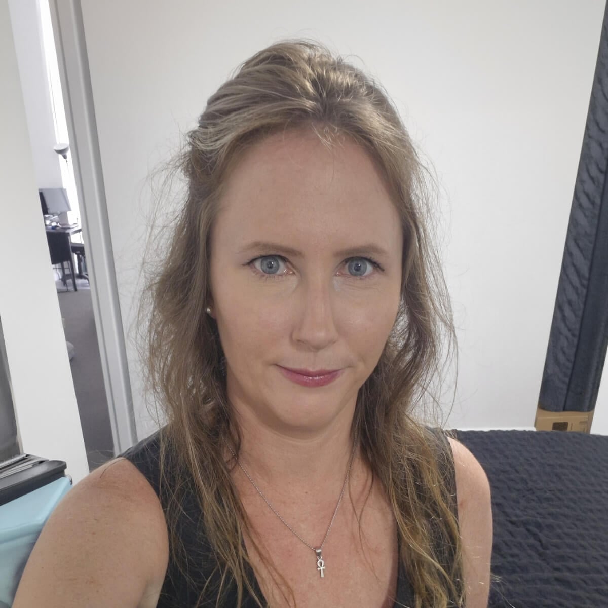 A woman with long hair and blue eyes is looking at the camera, wearing a black top and a necklace.