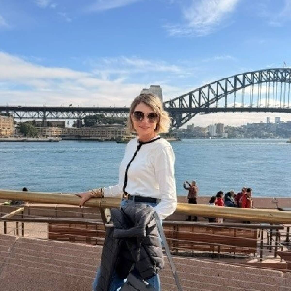 A woman in a white top and sunglasses stands by the Sydney Harbour, with the Sydney Harbour Bridge in the background.