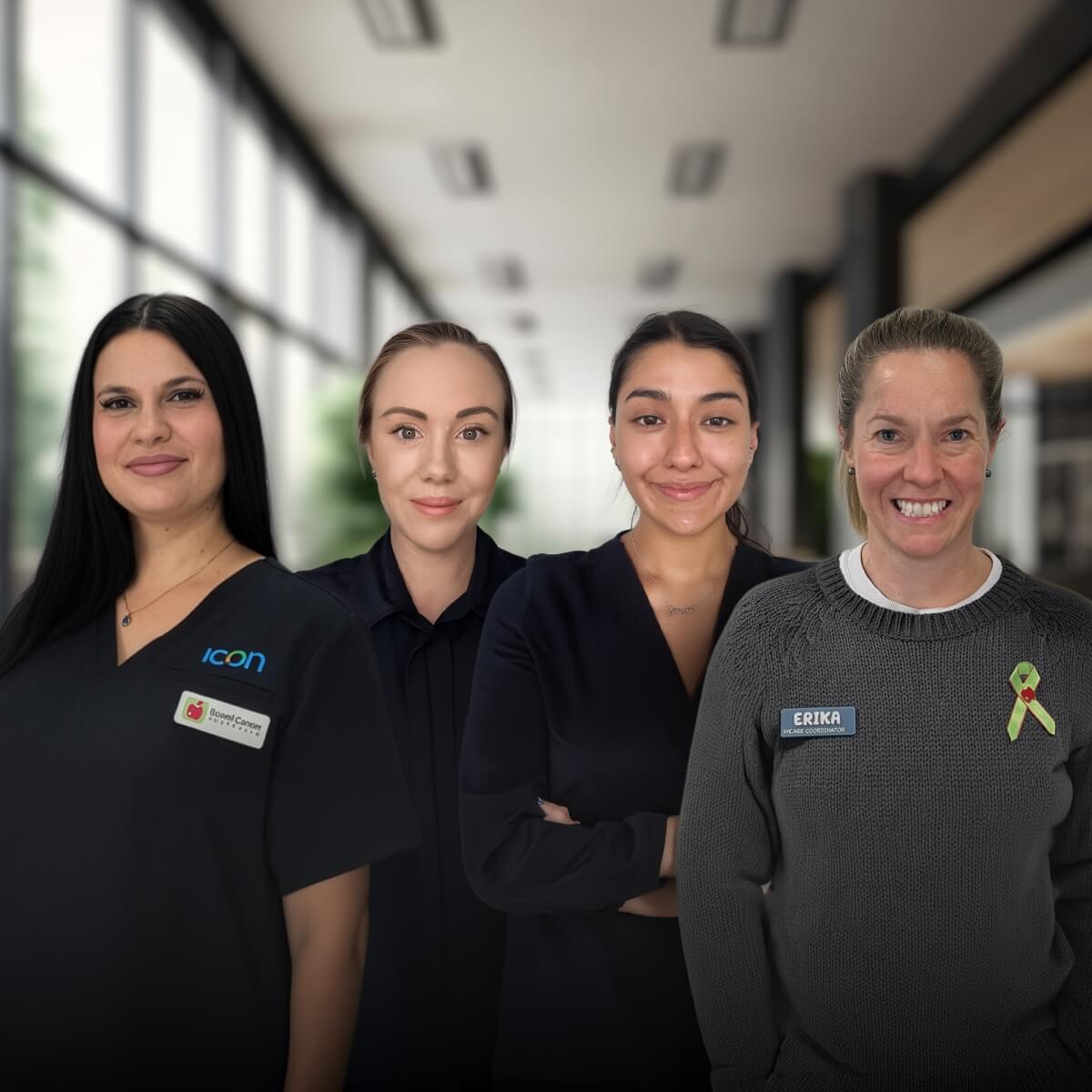 Four women stand together, smiling, in a well-lit indoor space, wearing professional attire and name tags.