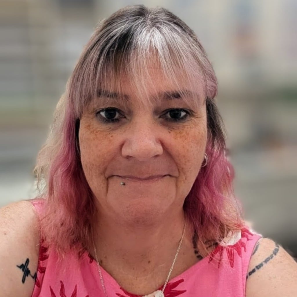 A woman with pink hair and a floral top smiles at the camera, set against a blurred background.