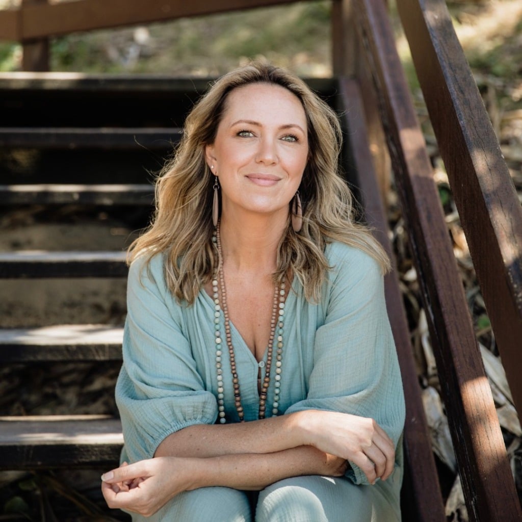 A woman in a light blue outfit sits on wooden stairs, smiling softly, with long hair and beaded jewelry.