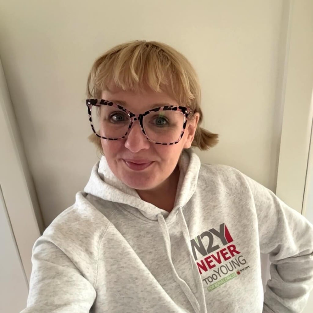 A smiling person wearing a gray hoodie with "Never Too Young" printed on it, promoting early-onset bowel cancer advocacy.