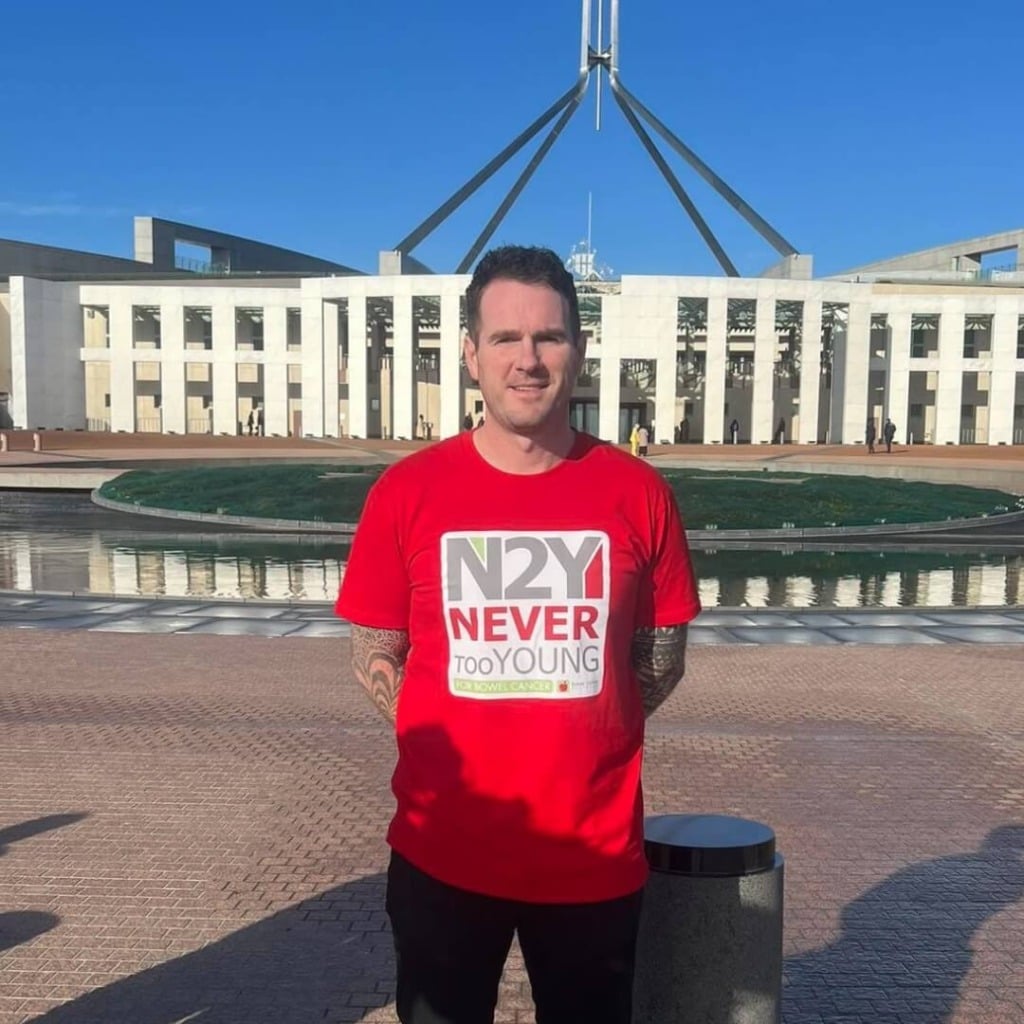 A person in a red "N2Y NEVER too YOUNG for bowel cancer" shirt stands in front of the Australian Parliament House.