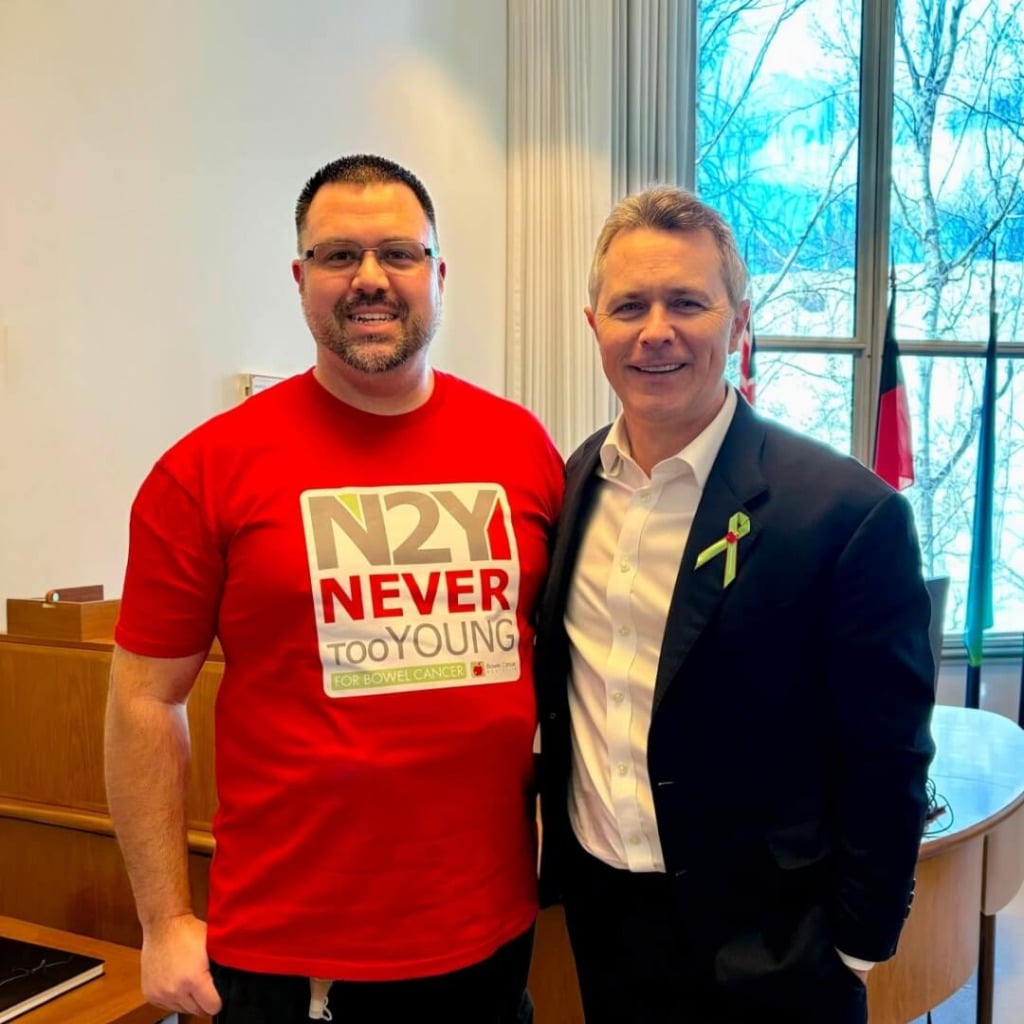 Two men pose together in an office setting, promoting early-onset bowel cancer advocacy with a "Never Too Young" shirt.