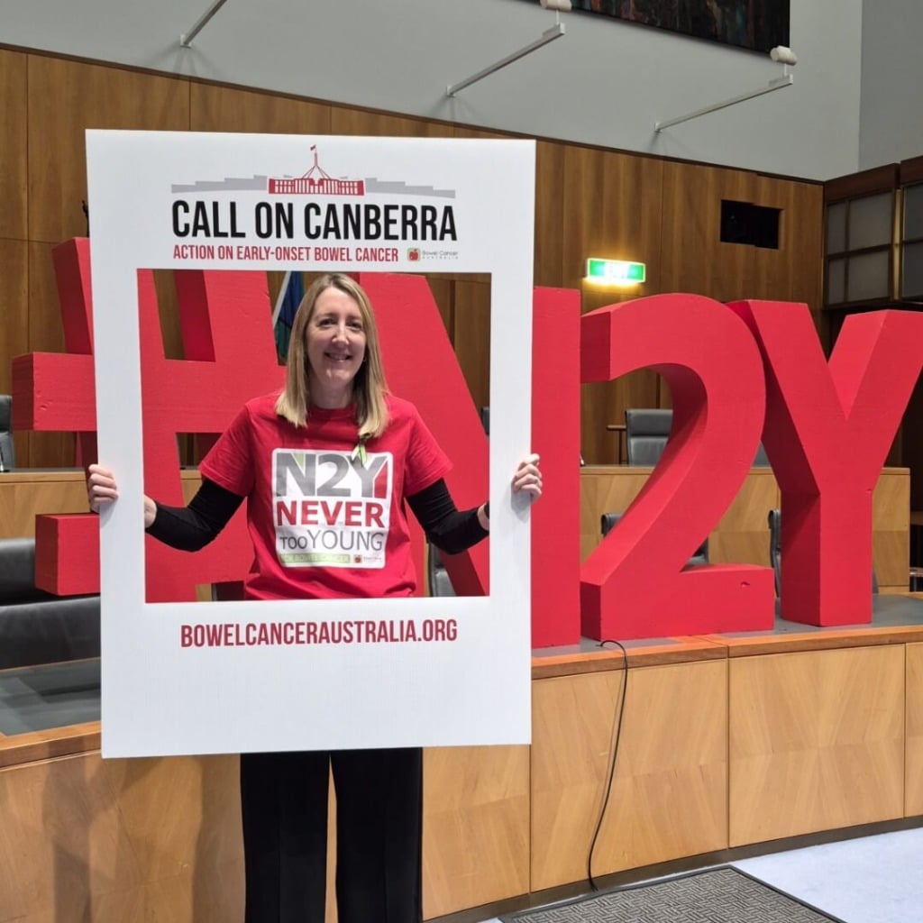 A woman in a red "Never Too Young" shirt poses in a frame with "Call on Canberra" and a large hashtag, advocating for bowel cancer awareness.
