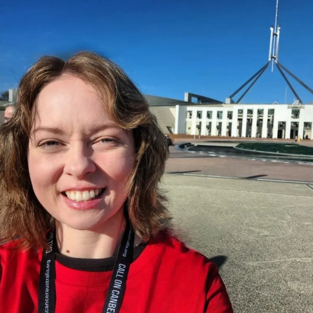 A smiling person in a red shirt stands in front of the Australian Parliament House, promoting early-onset bowel cancer advocacy.