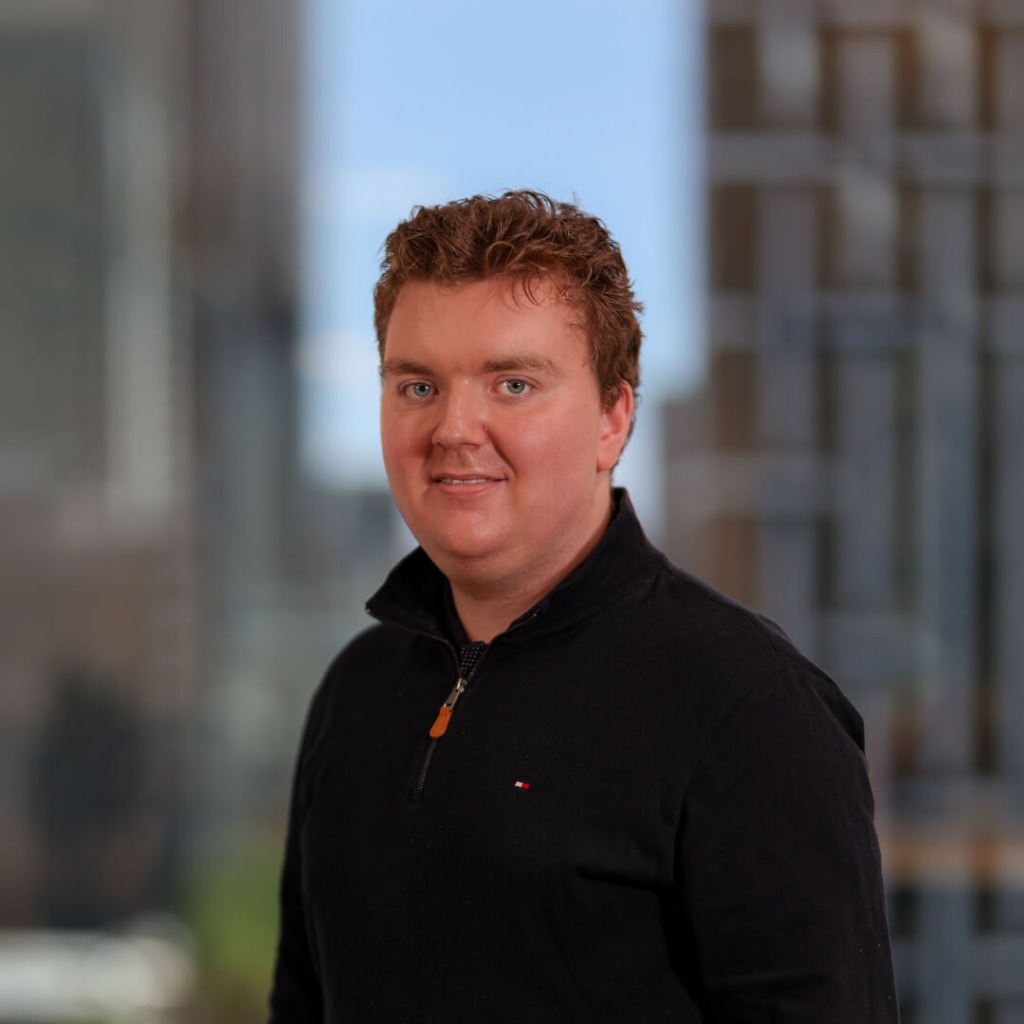 A young man with short brown hair smiles, wearing a black zip-up sweater, with a blurred city background.