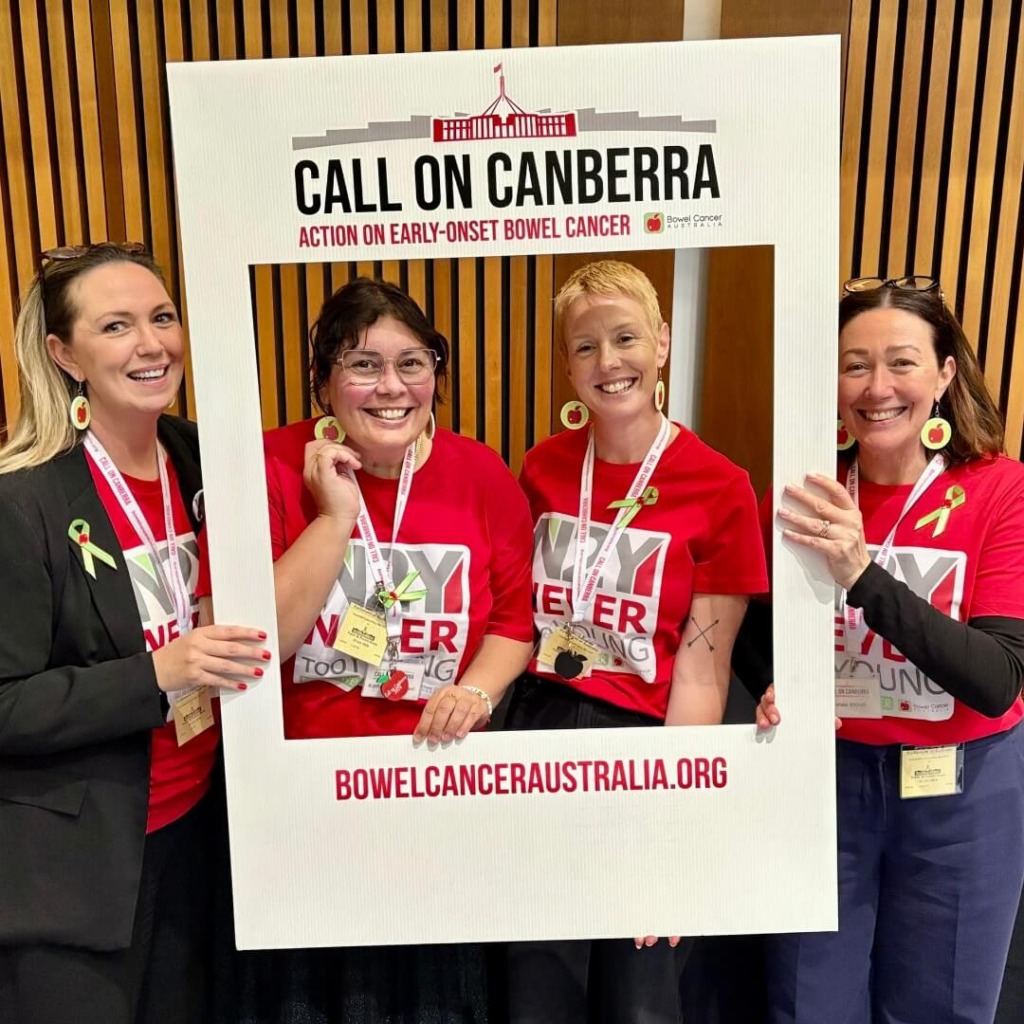 Four women hold a frame that reads "Call on Canberra" at an early-onset bowel cancer advocacy event, smiling and wearing red shirts.