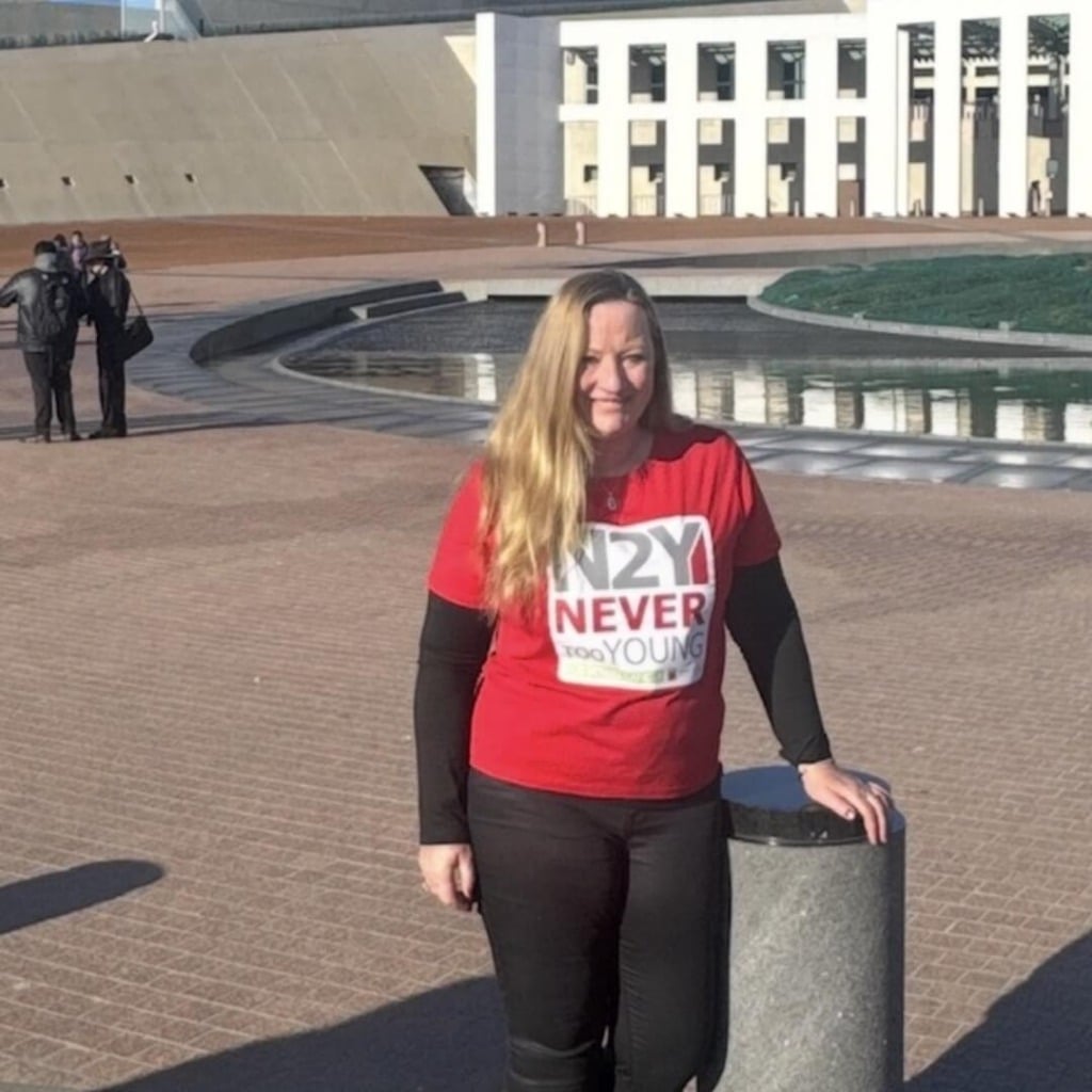 A woman in a red shirt stands near a water feature at Parliament House, promoting early-onset bowel cancer advocacy.