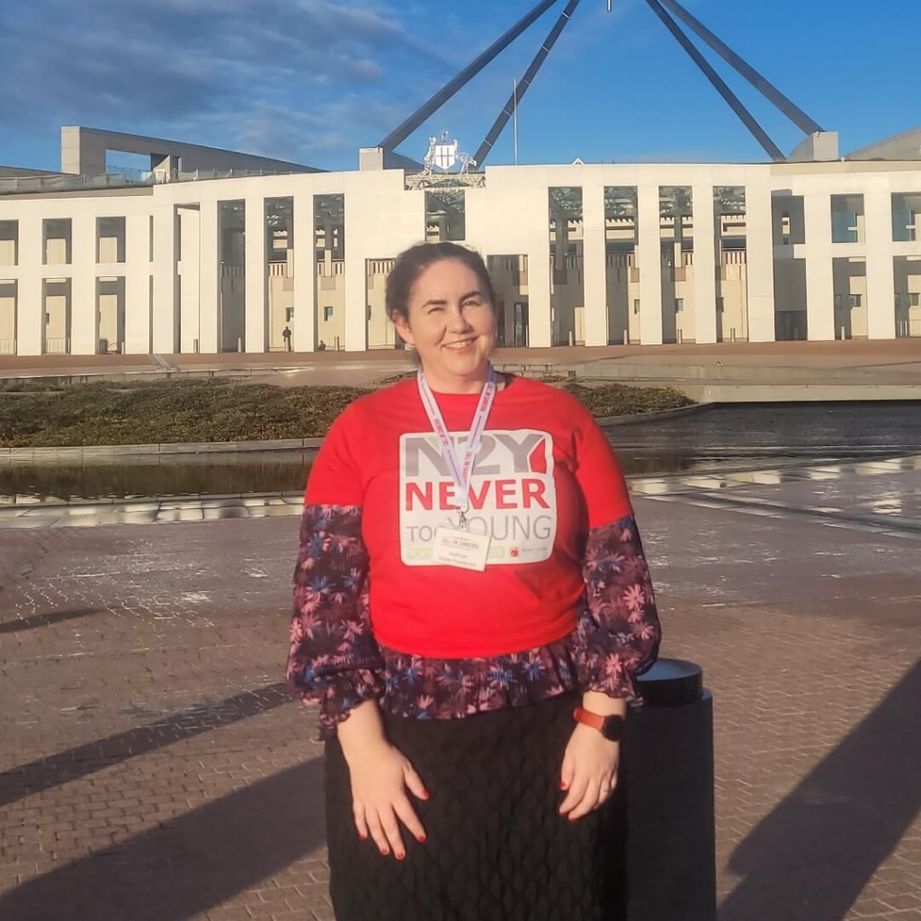 A woman in a red "Never Too Young" shirt stands in front of Parliament House, promoting early-onset bowel cancer advocacy.