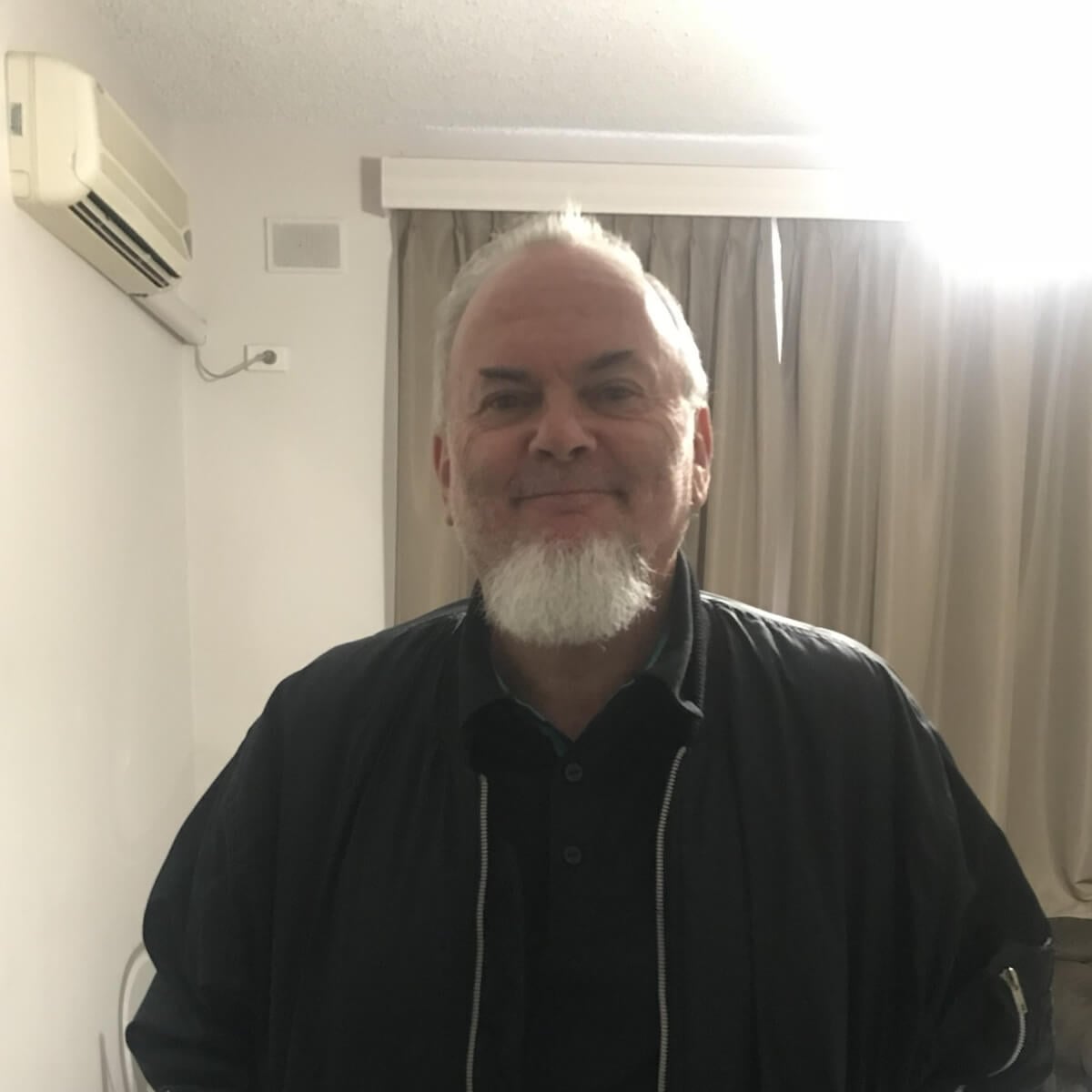 A smiling man with a beard stands in a room with light curtains and an air conditioning unit on the wall.