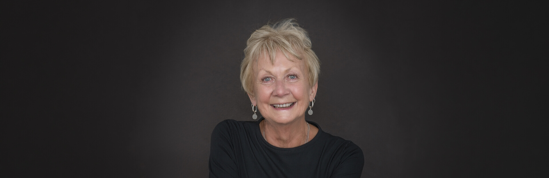 A smiling older woman with short blonde hair, wearing a black top, against a dark background.
