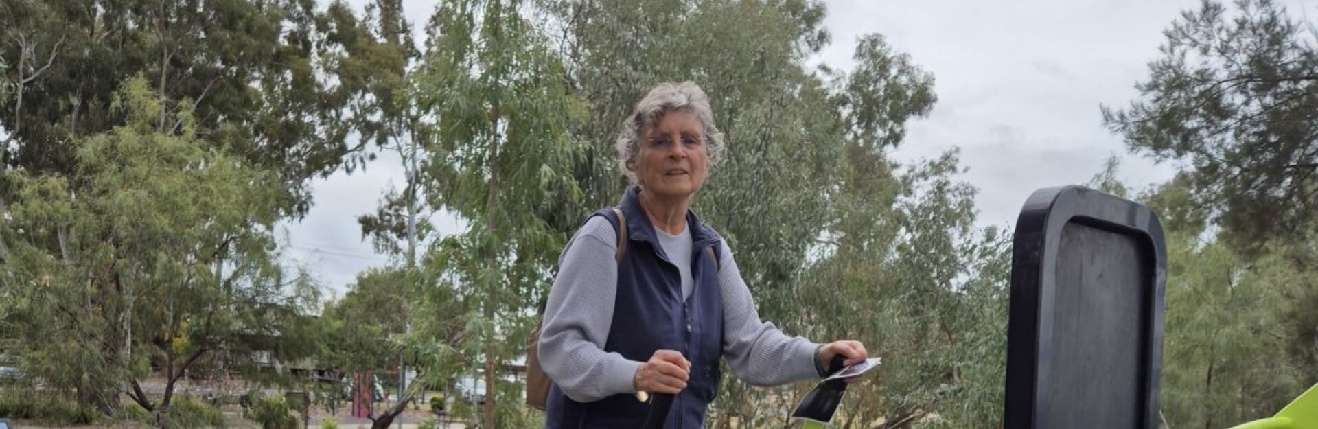 An older woman stands outdoors, smiling, with trees in the background and holding a device in her hand.