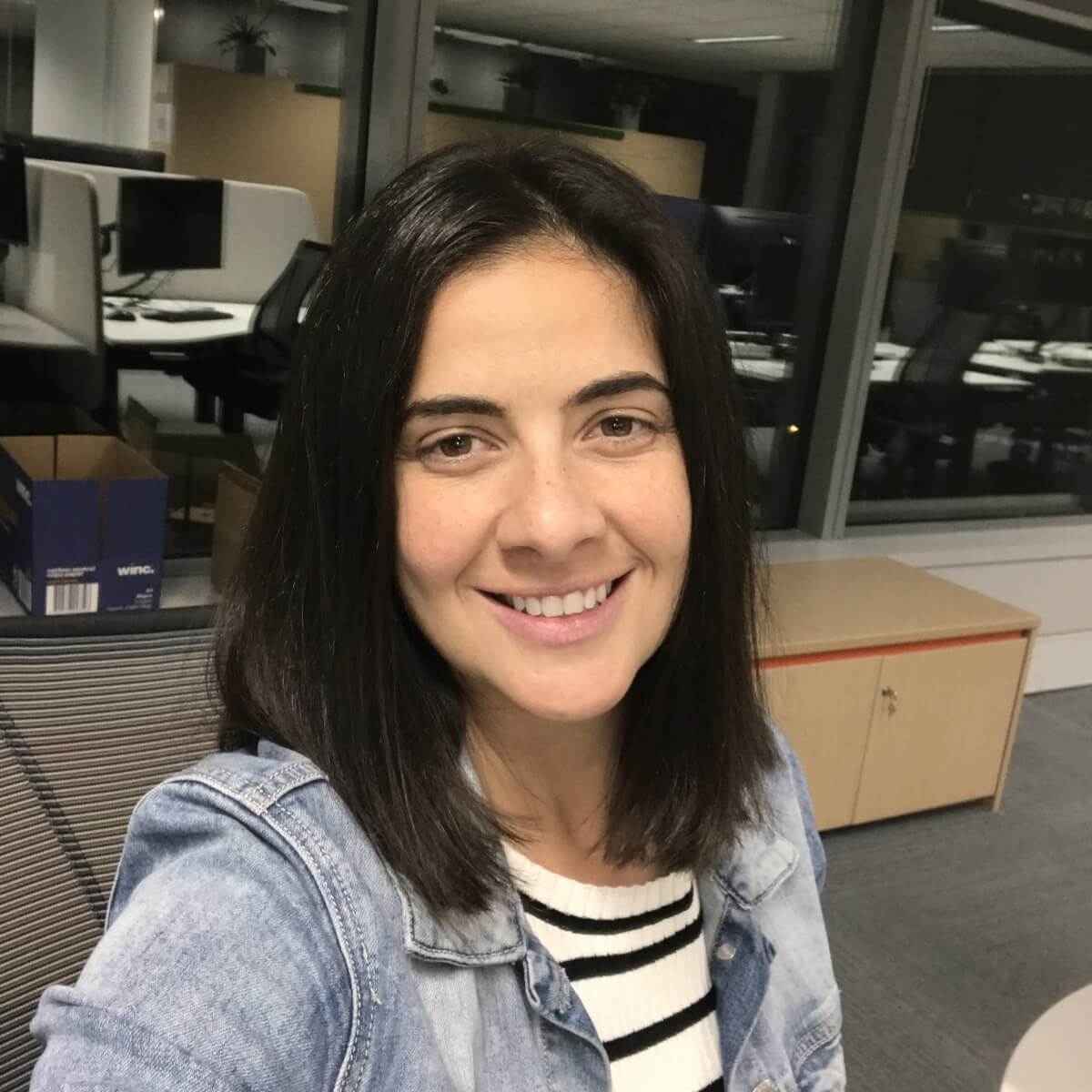 A woman with long dark hair smiles at the camera in an office setting with computers in the background.