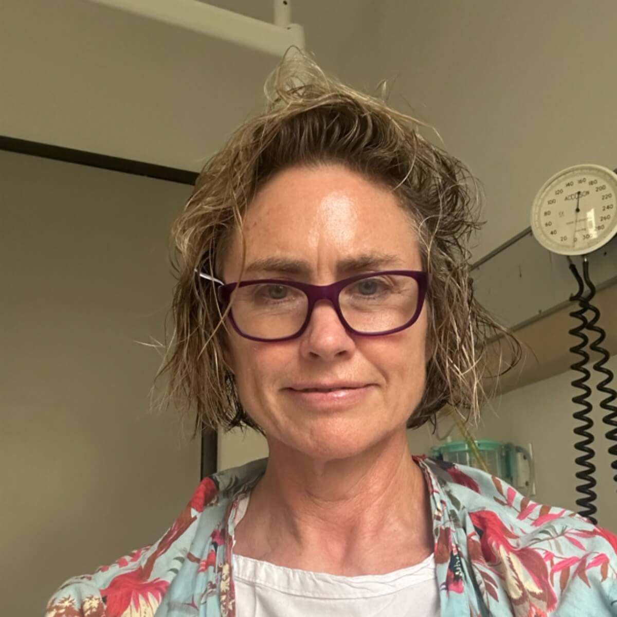 A woman with short, curly hair and purple glasses smiles in a medical setting with equipment in the background.