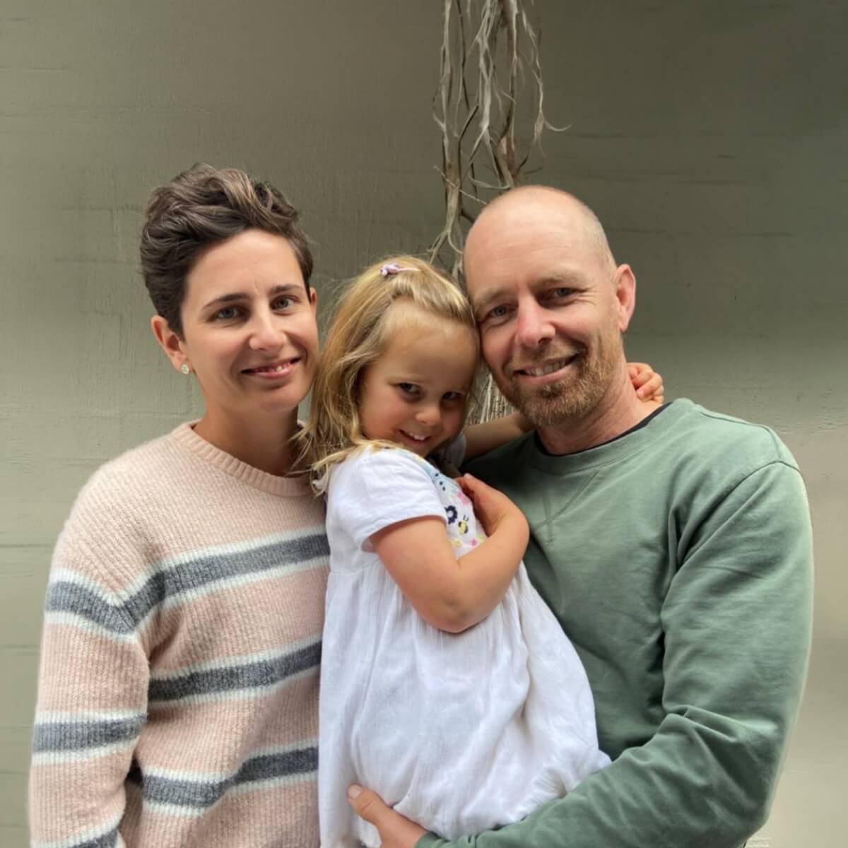 A man and woman pose together with a young girl, smiling, in front of a neutral background.