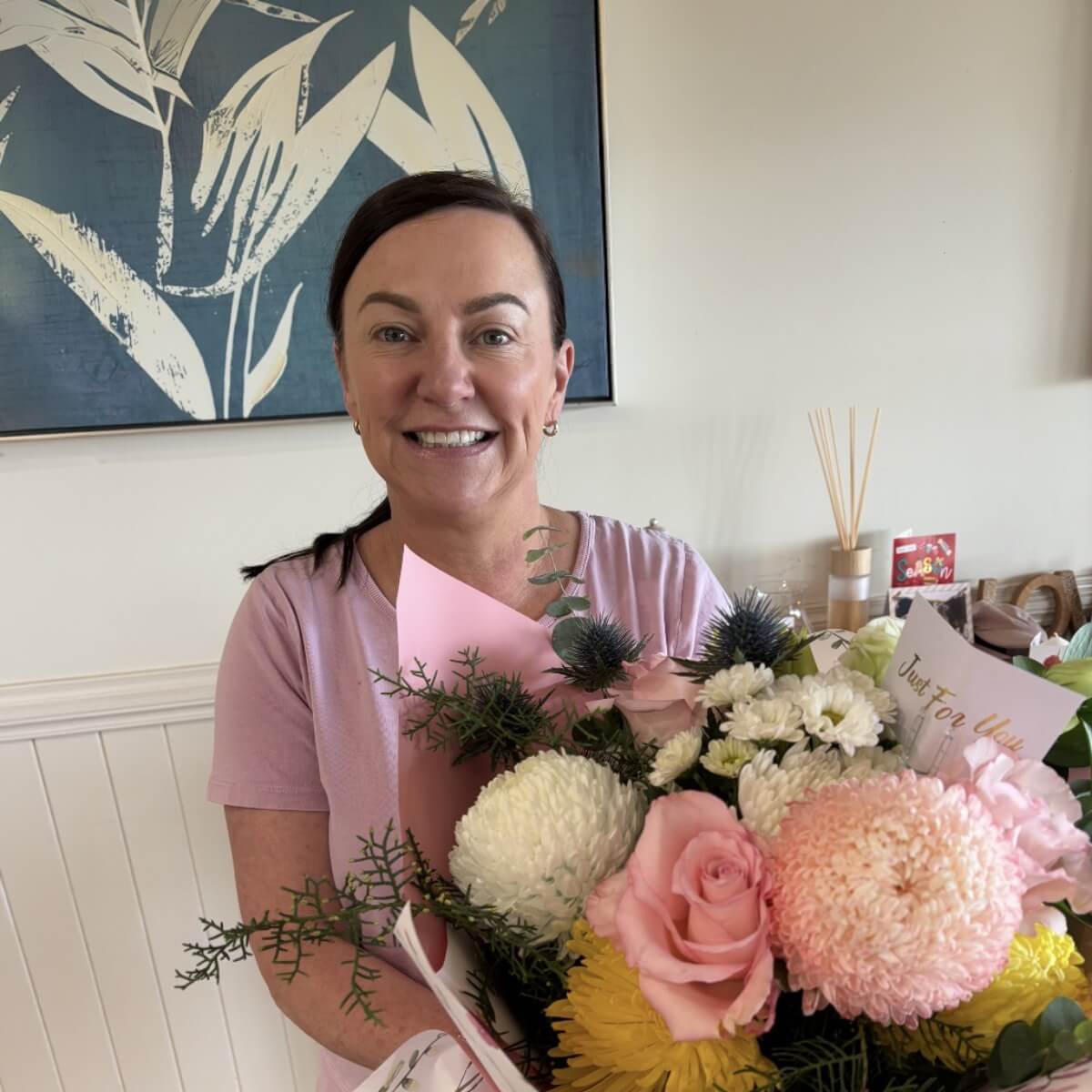 A woman in a pink shirt smiles while holding a large bouquet of colorful flowers with a card.