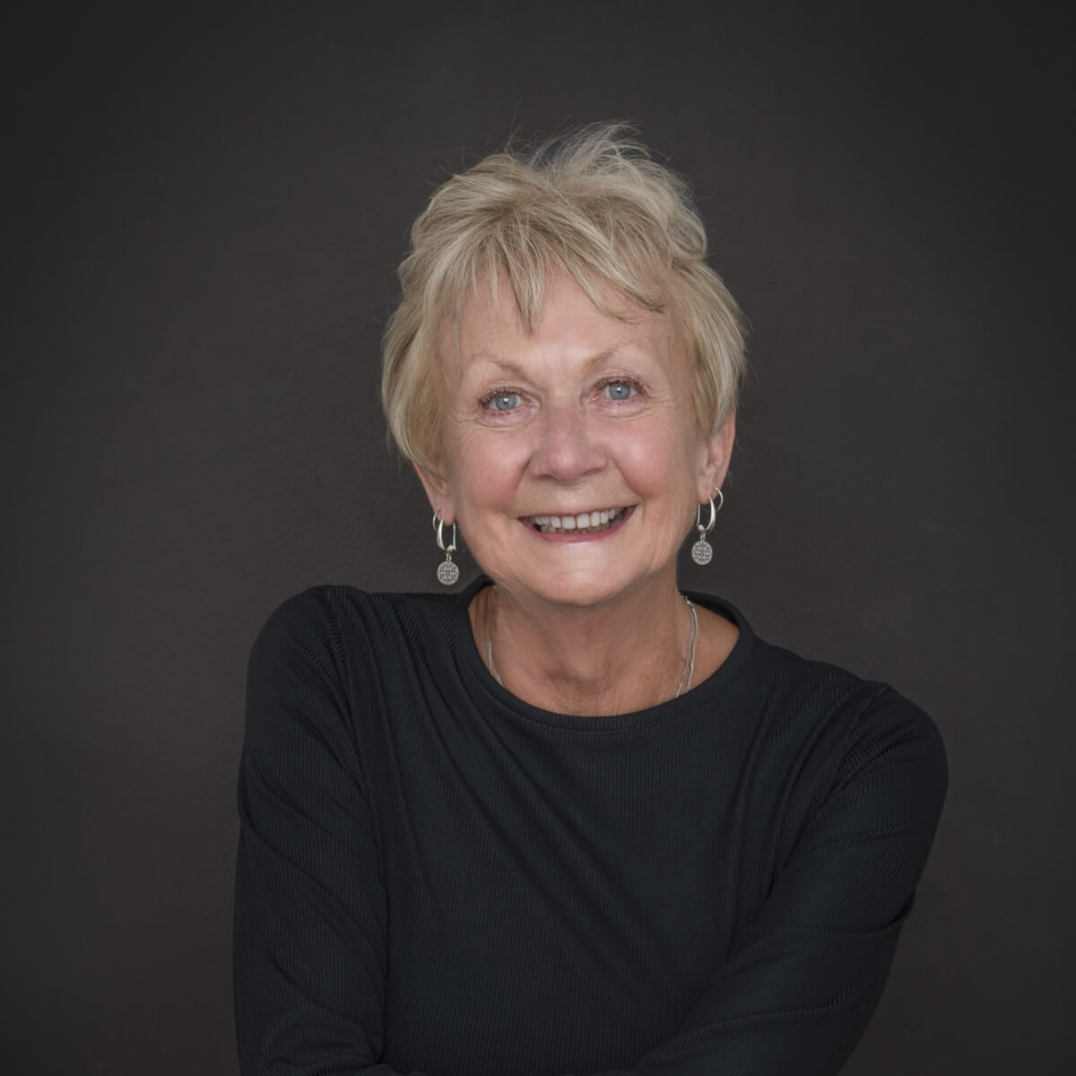 A smiling older woman with short blonde hair, wearing a black top and earrings, poses against a dark background.