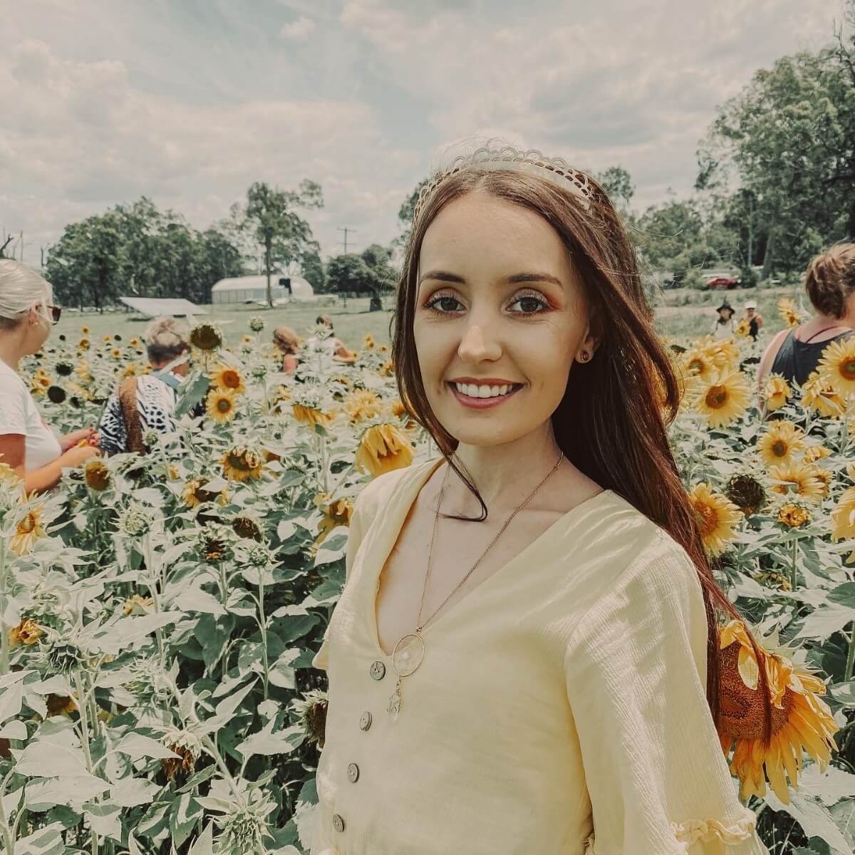 A young woman in a yellow top smiles in a sunflower field, surrounded by other people and bright sunflowers.