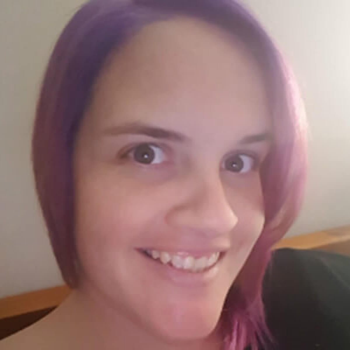 A woman with purple hair smiles at the camera, sitting against a light background.