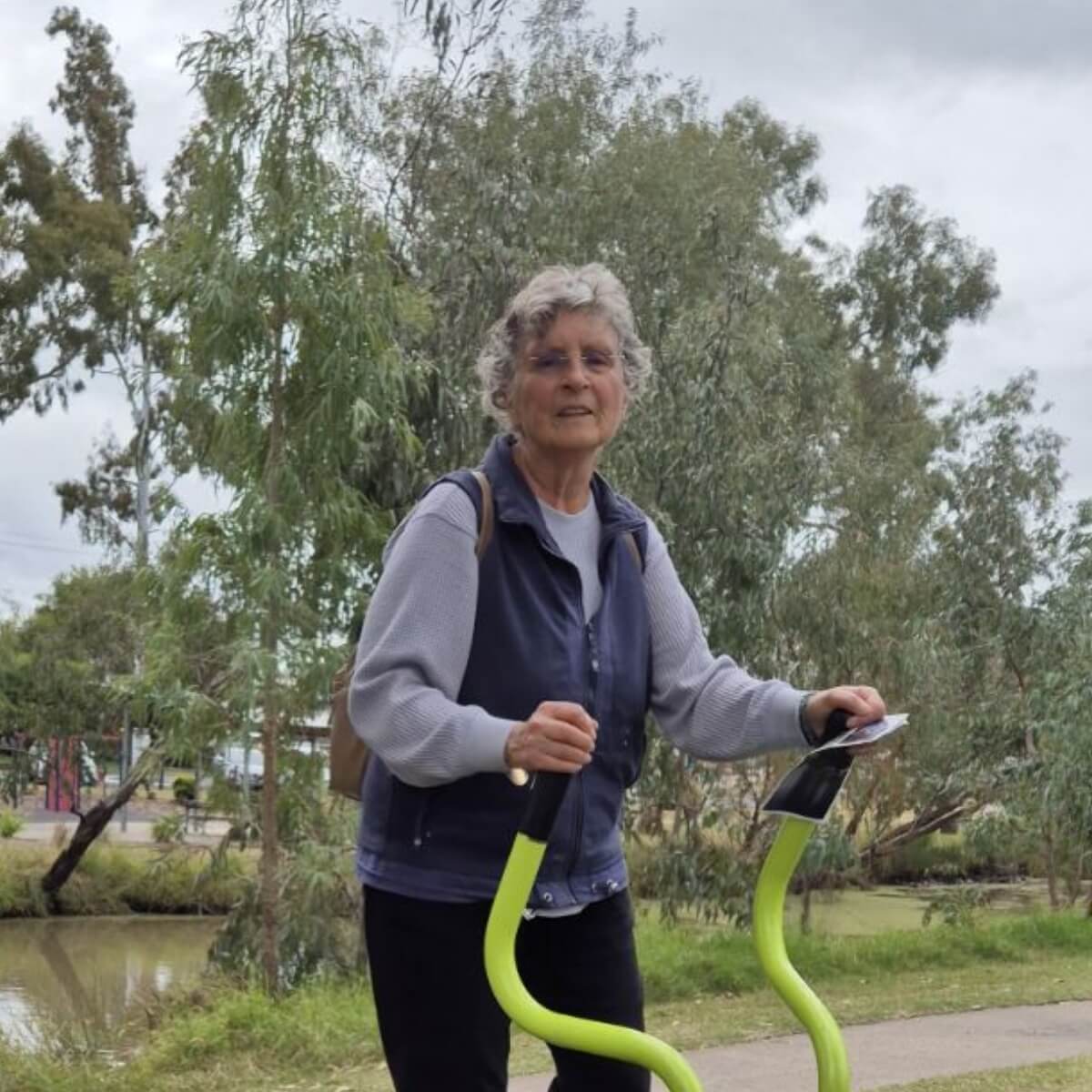 An older woman with curly hair stands by exercise equipment in a park, surrounded by trees and a cloudy sky.