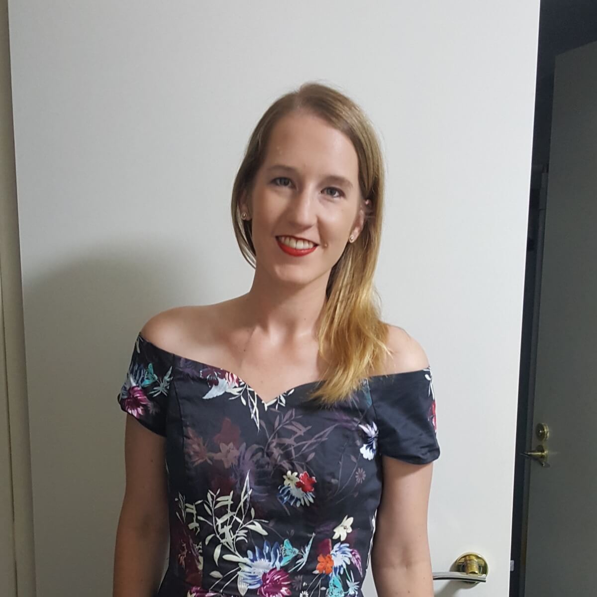 A woman with long blonde hair wearing a floral off-shoulder dress smiles in front of a white wall.