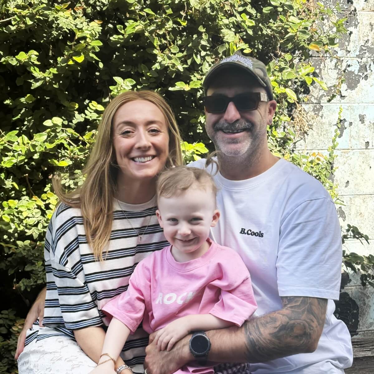 A smiling woman and man sit with a young boy in front of greenery, all looking happy and relaxed.