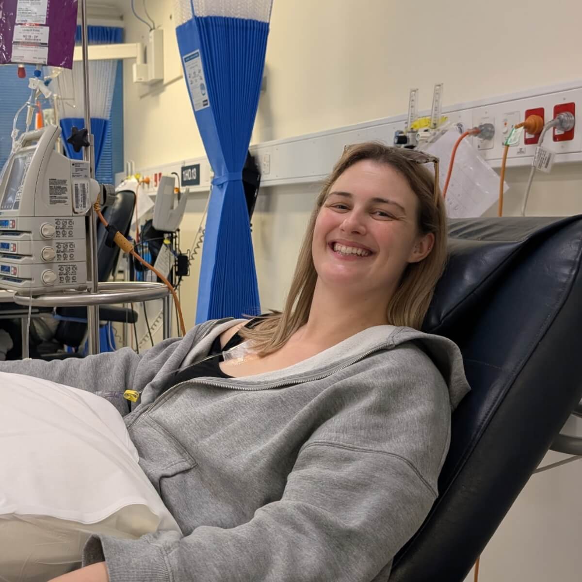 A smiling woman in a gray hoodie sits in a medical chair, with medical equipment visible in a clinic setting.