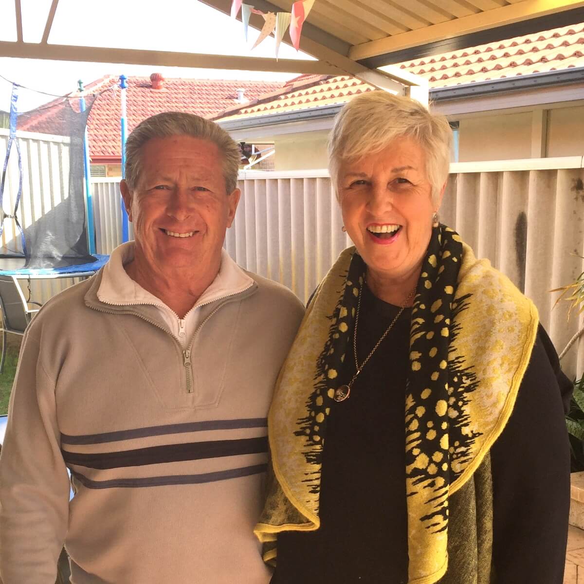 A smiling man and woman pose together outdoors, with a trampoline and house in the background.