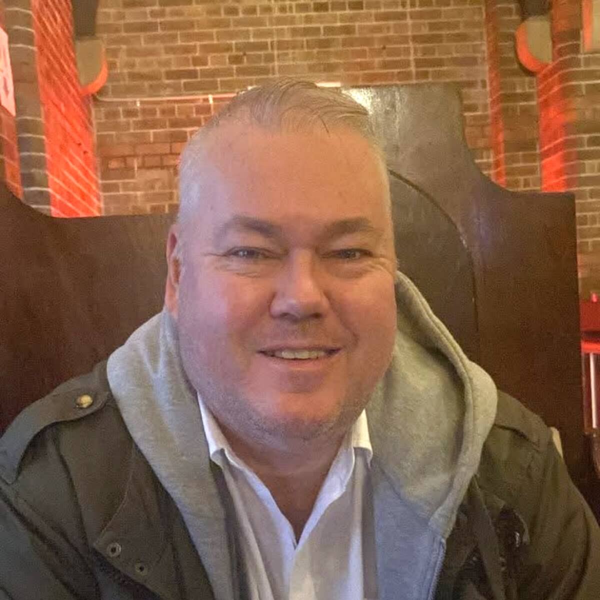 A smiling man with short hair wearing a gray hoodie and jacket, seated in a cozy, brick-walled environment.