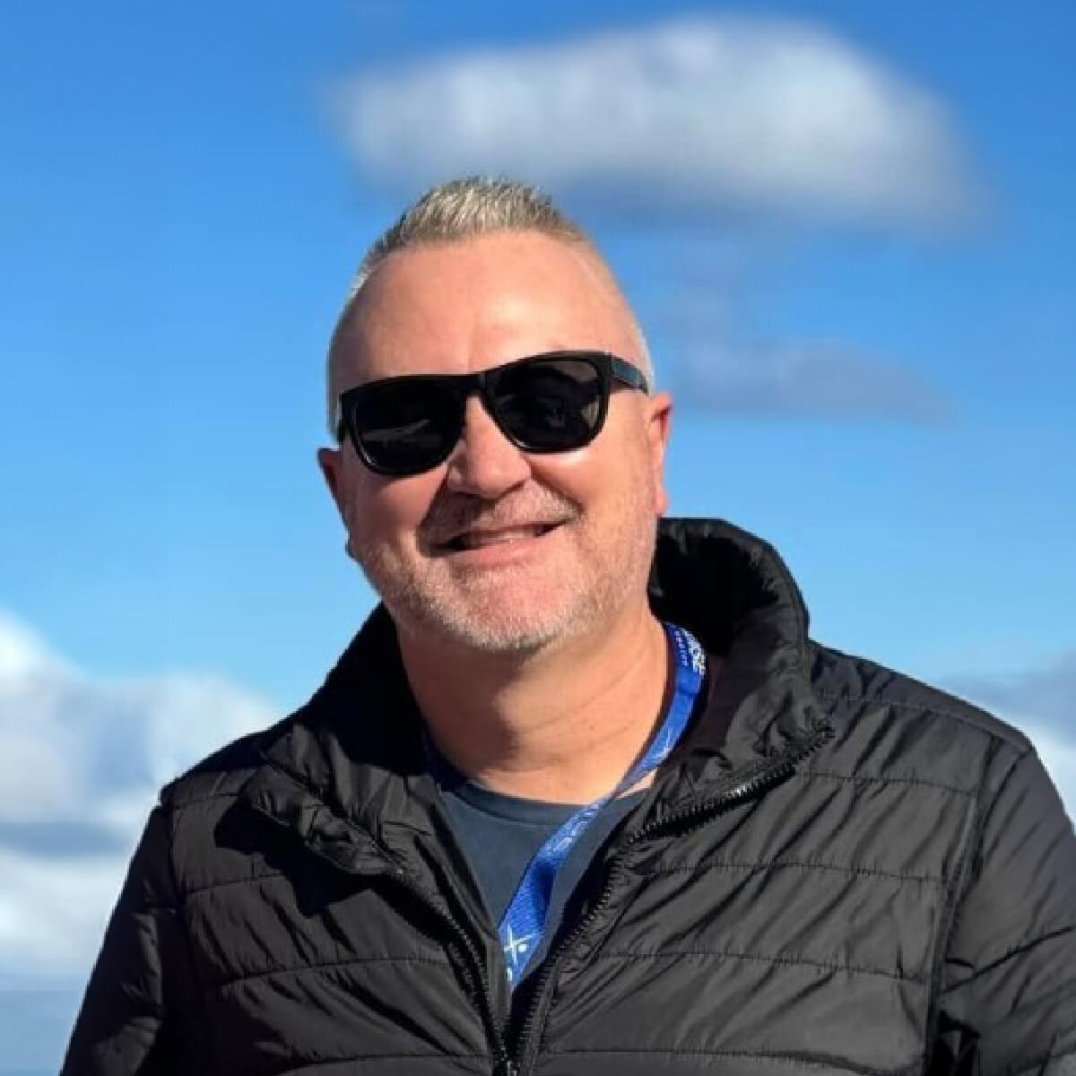 A smiling man with short, light hair and sunglasses stands outdoors against a blue sky with clouds.