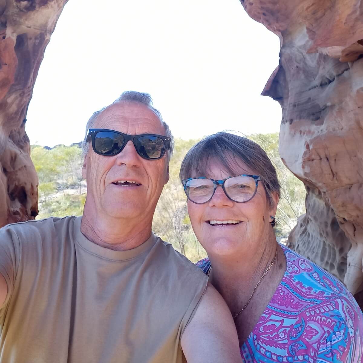 A man and woman take a selfie in a rocky outdoor setting, both smiling, wearing sunglasses, with greenery in the background.