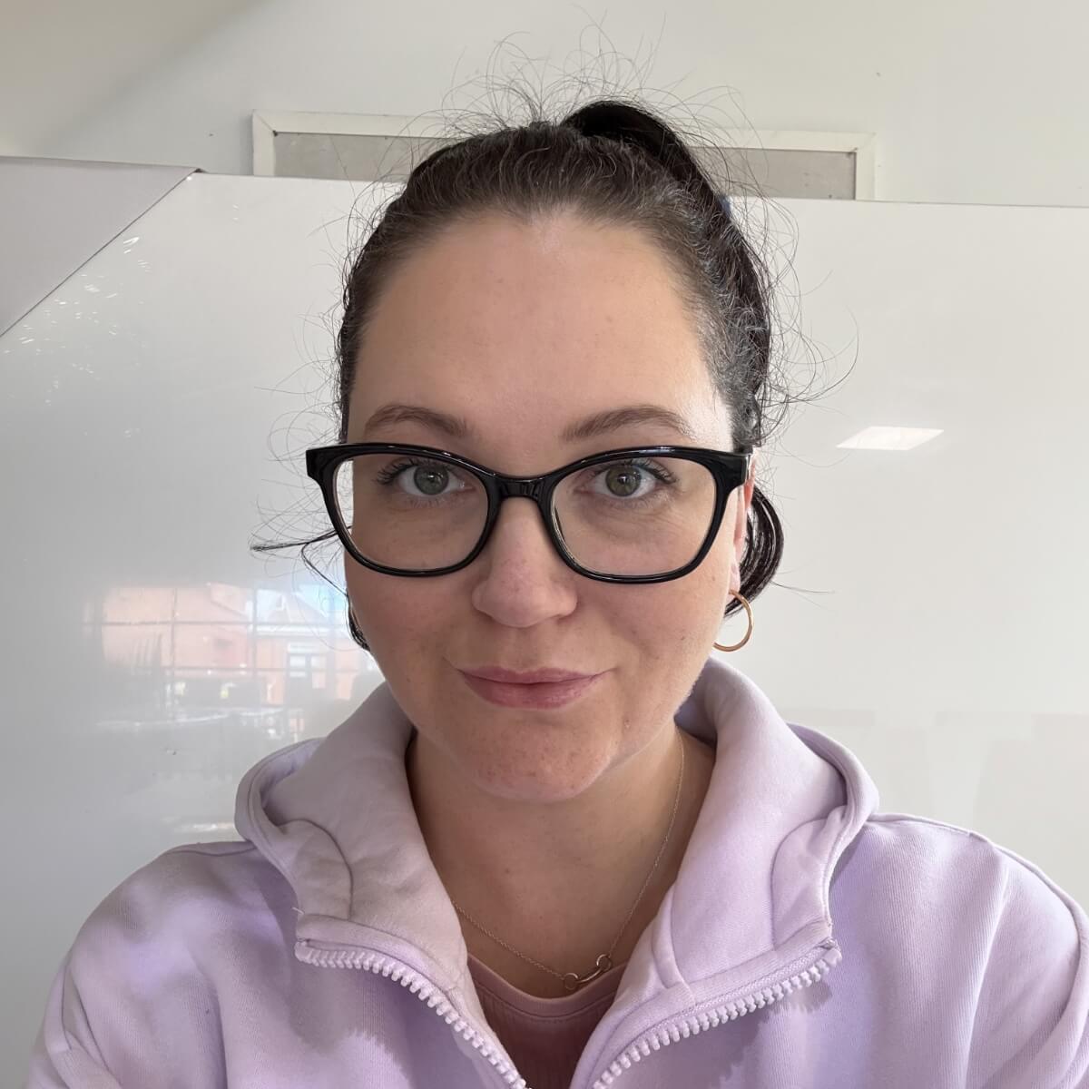 A woman with glasses and a light purple sweatshirt poses for a selfie against a bright background.
