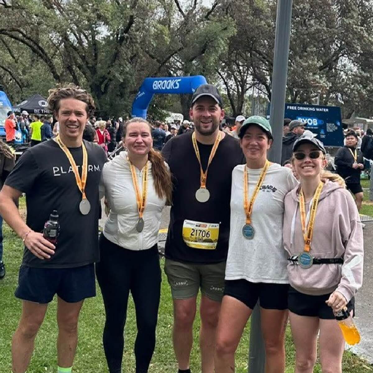Five smiling individuals stand together, each wearing a medal, in a park setting with trees and event banners in the background.