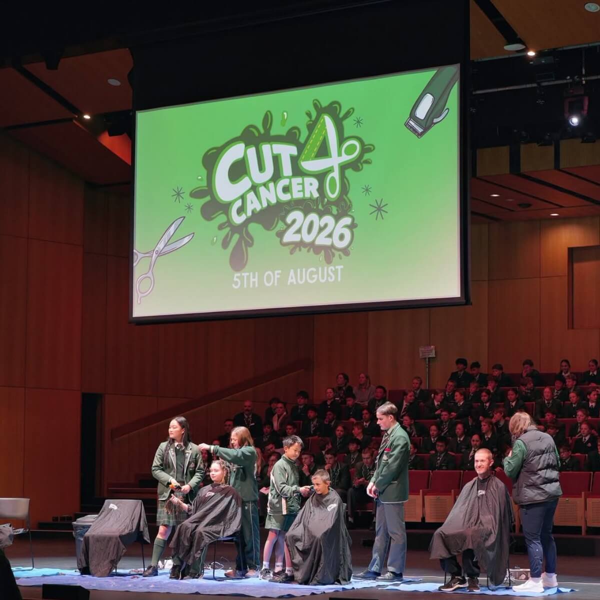 A stage with students getting haircuts, a banner for "Cut 4 Cancer 2026" in the background, audience seated.