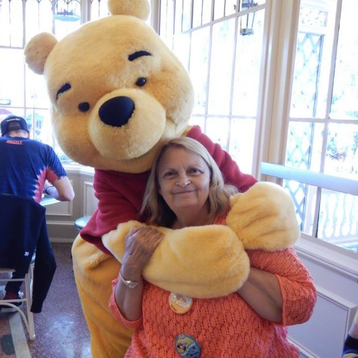 A woman with long hair smiles while hugging a large Winnie the Pooh character in a bright, sunny indoor setting.