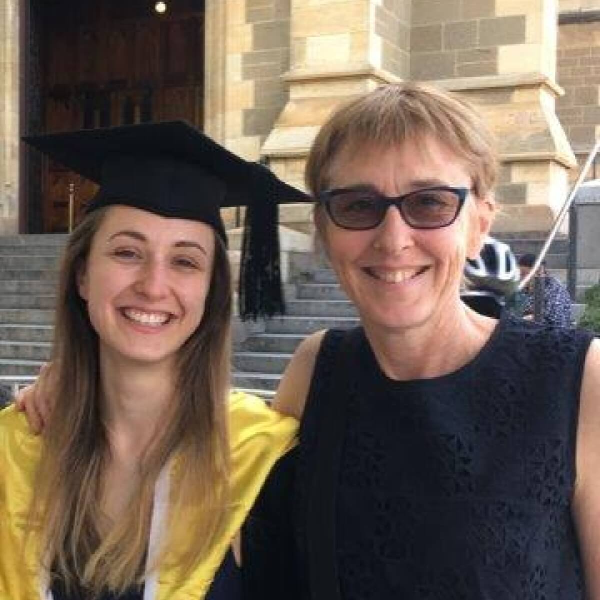 A smiling graduate in a cap and gown stands next to a woman, both in front of a building with stone steps.