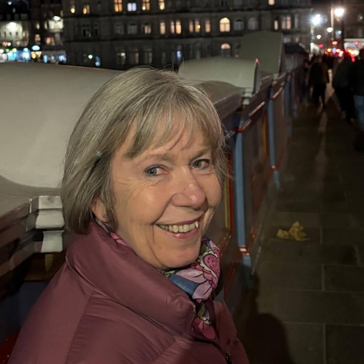 A smiling older woman with short gray hair, wearing a pink jacket and scarf, stands on a lit street at night.