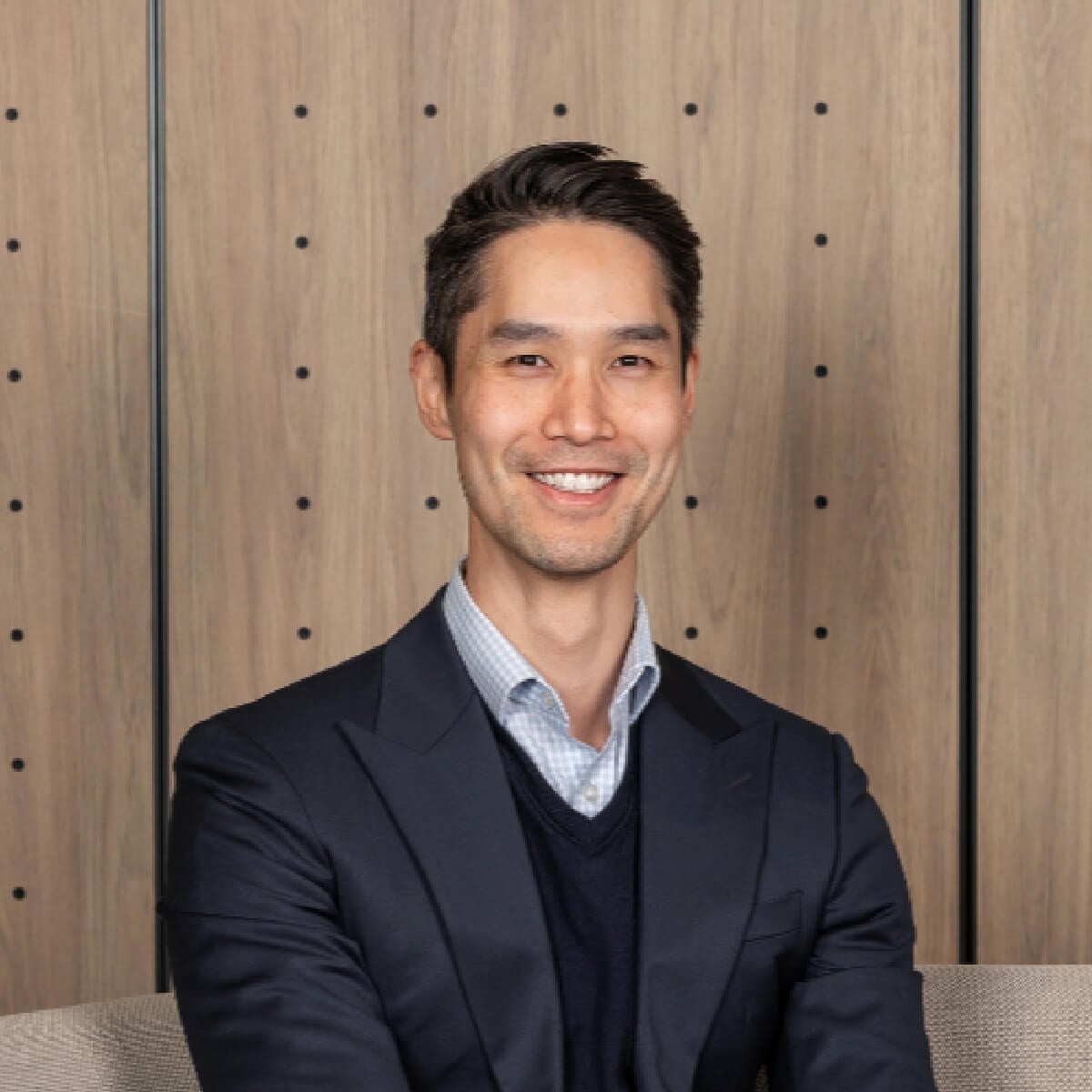 A smiling man in a suit poses against a wooden wall with small holes, sitting on a beige couch.