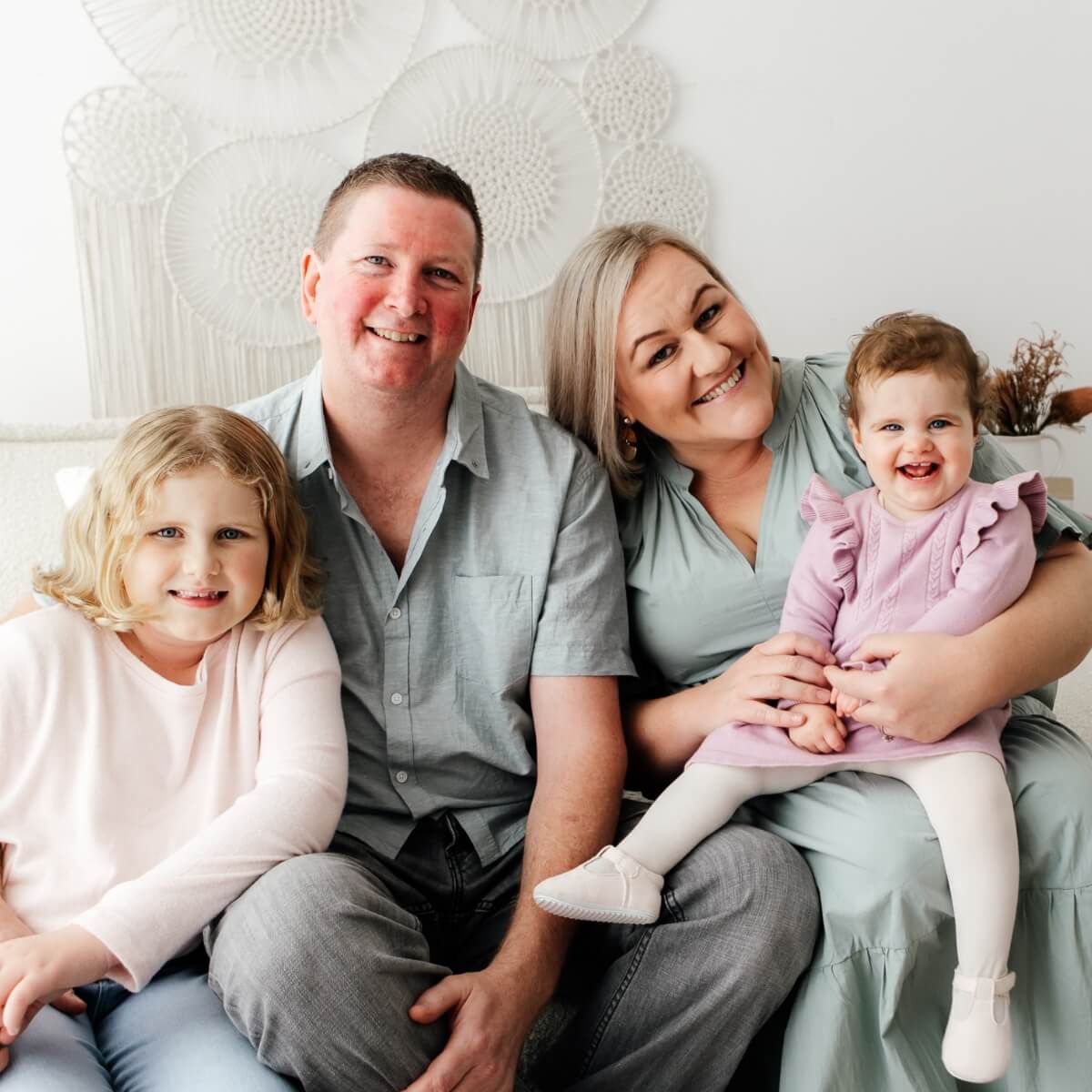 A family of four sits together on a couch, smiling. A woman holds a toddler, while a girl sits beside a man.