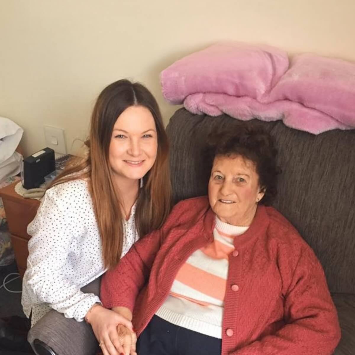 A young woman and an older woman sit together on a couch, smiling, with pink pillows in the background.