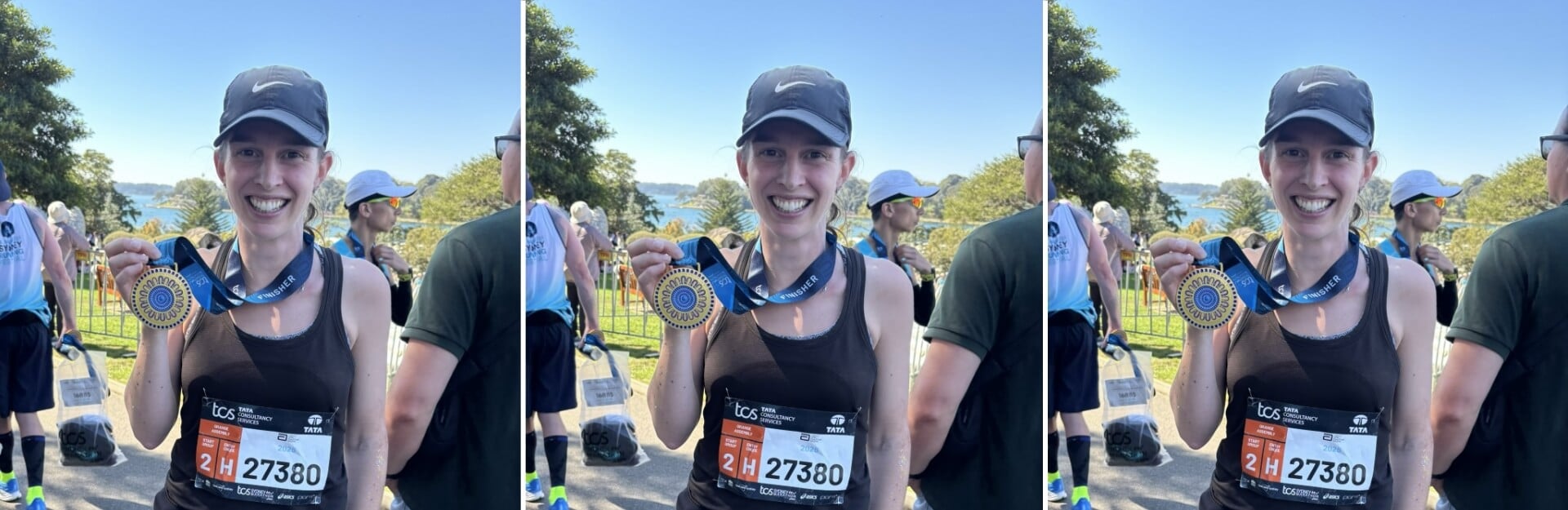 A smiling woman in a black tank top holds a marathon finisher medal, surrounded by other participants and a scenic background.